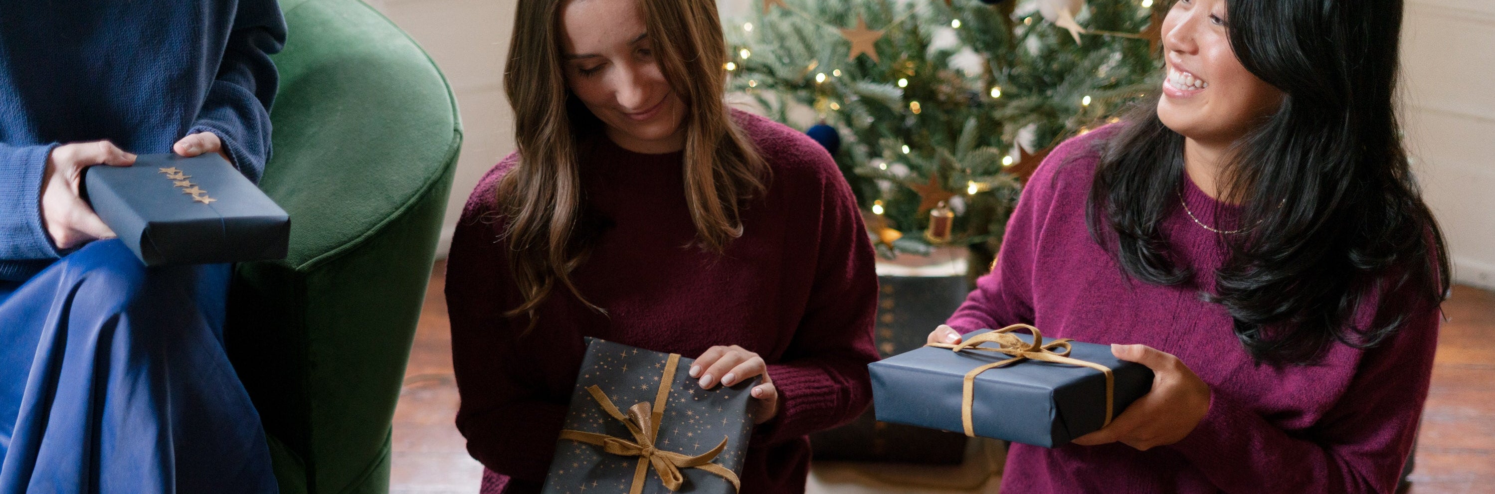 Three women sitting on the floor holding wrapped gifts in front of a Christmas tree.