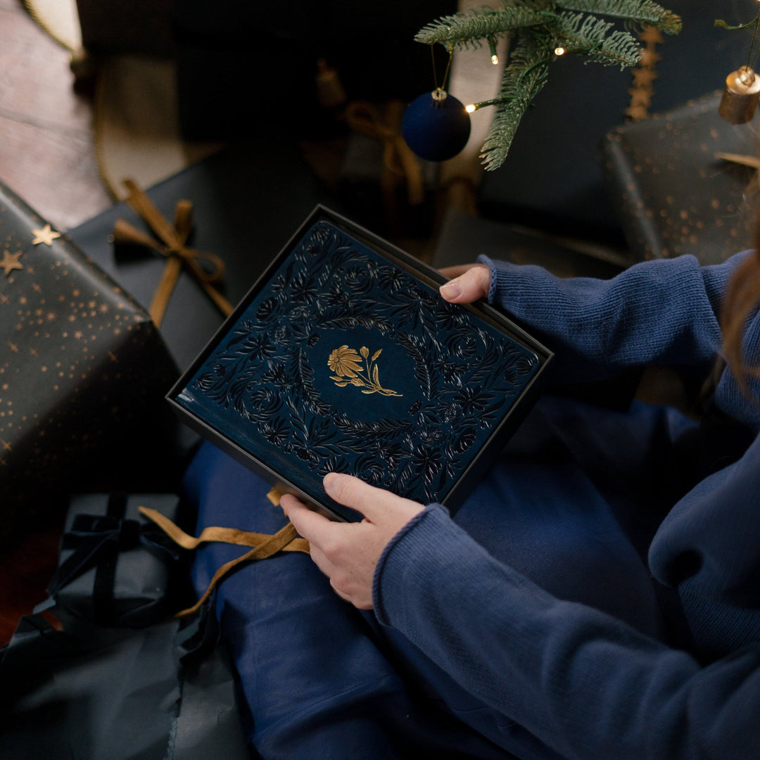 Person holding a dark blue gift box with gold accents in a festive setting with Christmas trees and wrapped gifts.