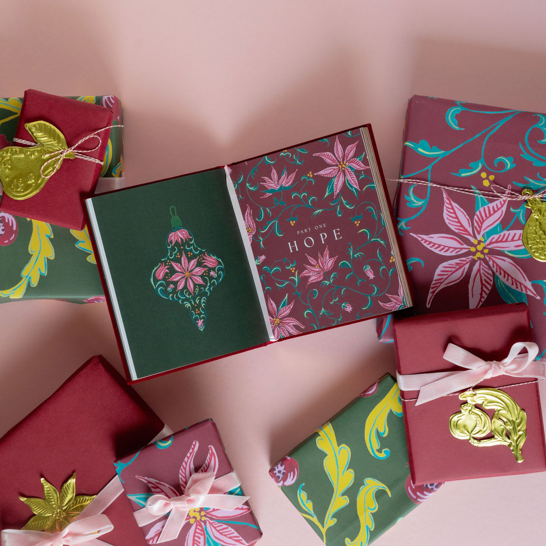 A styled flat-lay of Christmas-themed wrapping paper and gift boxes on a solid pink background. The wrapping paper features various festive patterns including floral motifs, gold foil ornaments, and green and burgundy backgrounds.