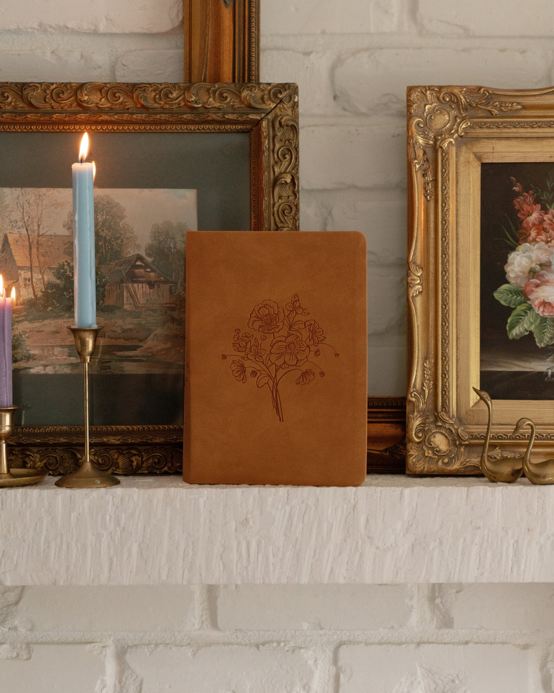 Decorative shelf with candles, a leather notebook, and framed artwork against a white brick wall.