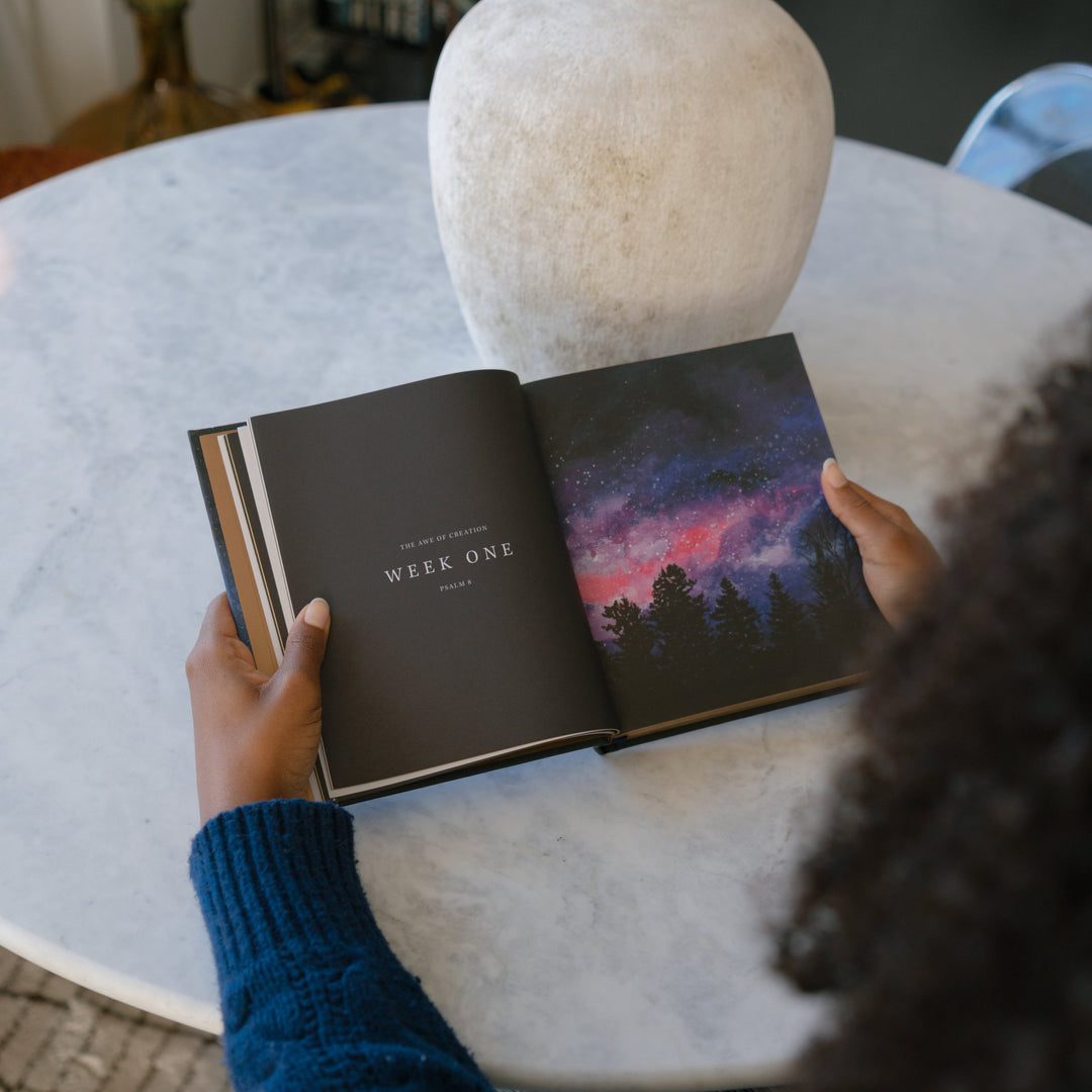 Person holding an open book with a cover featuring a purple landscape on a marble table.